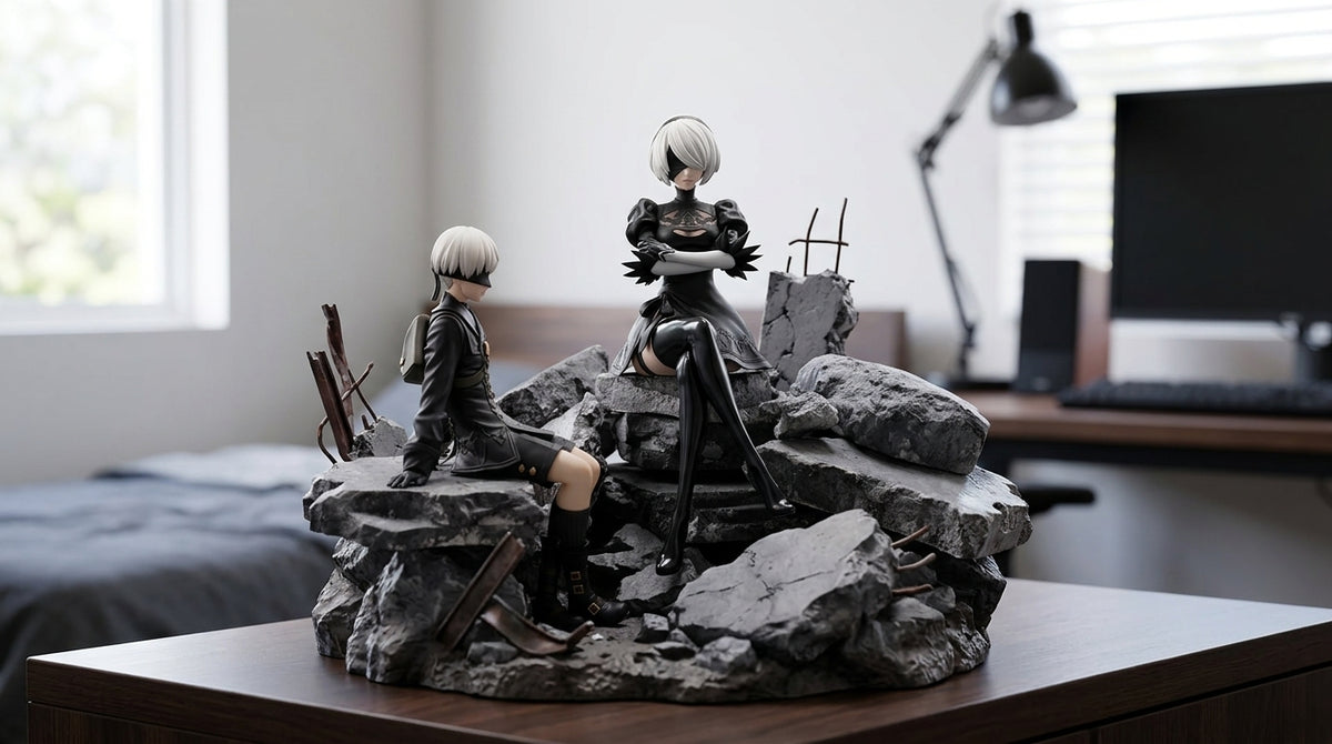 2B and 9S inspired figures displayed on rocky rubble diorama base on wooden desk with monitor and lamp visible in background gaming room setting
