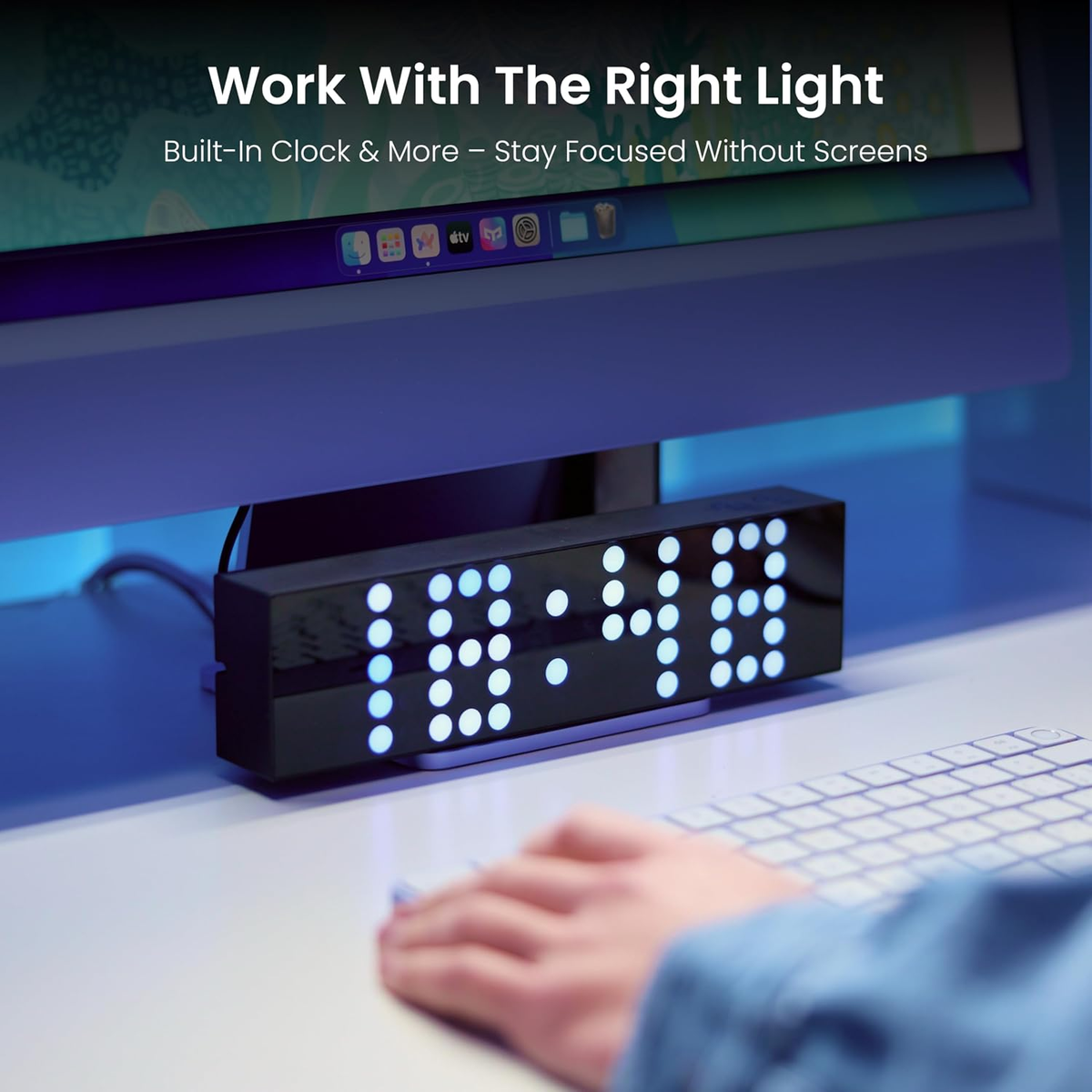 A black rectangular LED digital clock sits on a desk under a computer monitor, showing the time in glowing blue digits. A person's hand and arm are visible at the lower right, using a keyboard in a workspace setting.